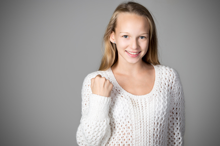 Portrait of happy beautiful casual caucasian girl wearing white knitted sweater, looking at camera with smile, showing winner gesture, studio image, gray background, focus on handの写真素材