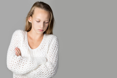 Portrait of unhappy beautiful casual caucasian girl wearing white knitted sweater, standing with crossed arms, looking down, studio image, gray background, copy spaceの写真素材