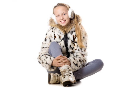 Portrait of happy funny beautiful casual teenage girl wearing winter coat and earmuffs, sitting cross-legged, looking at camera with cheerful expression, studio image, white backgroundの写真素材