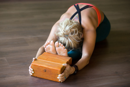 Sporty beautiful young blond woman in sportswear working out indoors, doing exercise for spine, shoulders, hamstrings, sitting in seated forward bend posture, focus on wooden bricksの写真素材