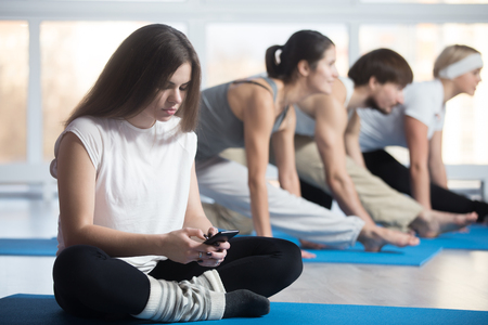 Tired or lazy woman sitting cross-legged, resting during practice in sports club, using phone, reading, sending or typing message, group of attractive fit young people working out on the backgroundの写真素材