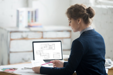 Beautiful young designer person wearing suit looking at paper with design drawing. Attractive model working at office desk with blueprints for new project. Interior shot. Back viewの写真素材