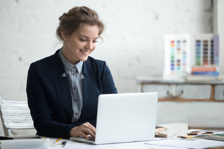 Portrait of beautiful happy smiling young designer woman working on laptop at office desk. Attractive cheerful model using computer. Typing indoorsの写真素材