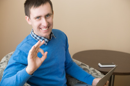 Portrait of casual attractive happy young man posing indoors, sitting in armchair with laptop,...
