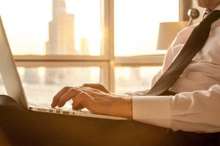 Arms of handsome young businessman wearing formal white shirt and tie typing on laptop sitting on the bed in modern room with sunny city view on background. Indoors. Close-upの写真素材