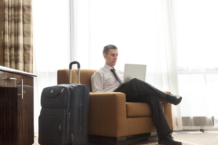 Portrait of handsome young business man wearing formal white shirt and black tie sitting in armchair in hotel room with packed luggage bag on the floor. Traveller working on laptopの写真素材
