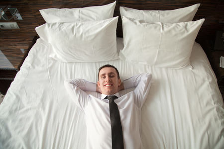 Portrait of young happy business man wearing white shirt and necktie lying on the bed in the hotel room with his hands behind head. View from aboveの写真素材