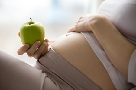 Close-up of torso of young pregnant model holding green apple. Future mom expecting baby eating fruits at home. Closeup of belly. Healthy eating conceptの写真素材