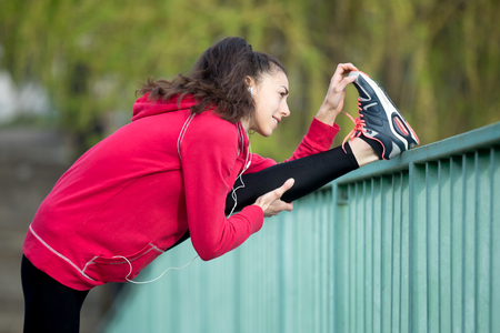 Portrait of sporty smiling woman doing hamstring stretch in park after jogging. Female athlete...