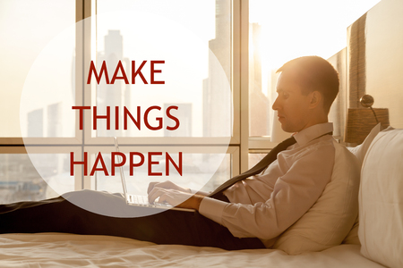 Portrait of handsome young businessman wearing white shirt and black tie sitting on bed with laptop in modern room with sunrise city view on background. Motivational text "Make things happen"の写真素材