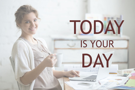 Portrait of beautiful happy smiling young designer woman sitting at home office desk with cup of coffee, looking at camera. Motivational phrase "Today is your day"の写真素材