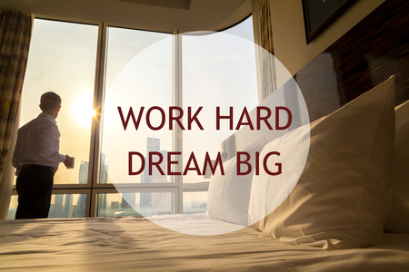 Maid-up bed in cozy room. Young businessman with coffee cup standing at window looking at city scenery on the background. Motivational text "Work hard Dream big"の写真素材