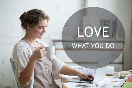 Portrait of beautiful happy smiling young designer woman sitting at home office desk with cup of coffee, working on laptop in loft interior. Motivational phrase "Love what you do"の写真素材