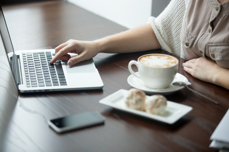 Hands of beautiful young woman sitting at table with cup of coffee and cakes and working on laptop. Attractive model using computer in cafe or office interior. Close-upの写真素材