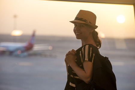 Young happy smiling woman in 20s waiting for flight in modern airport terminal building at sunrise or sunset, standing in front of window with view on takeoff groundの写真素材