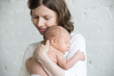 Portrait of young happy mother tenderly hugging sleeping adorable new born child and smiling. Funny newborn babe napping in mom arms. Mum and healthy little kid. Love, bonding, happy family concept.の写真素材