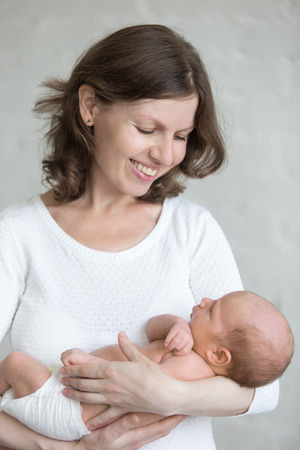 Portrait of young mother holding newborn cute babe in hands. Smiling mom looking at her newborn child with happiness. Healthy little kid after birth lying in parent arms. Happy family concept. Indoorsの写真素材