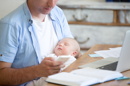 Casual business dad holding sleeping newborn babe while working in home office interior, holding smartphone and looking at screen. Young father using mobile phone and nursing new born child. Close-upの写真素材