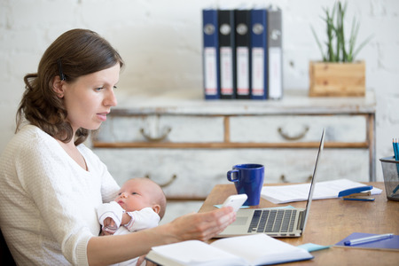 Portrait of young business mom holding her newborn cute babe while working in home office interior, looking at cellphone screen. Serious working mother using mobile phone and nursing new born childの写真素材