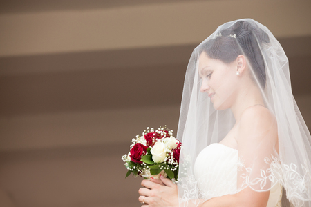 Side view portrait of young beautiful bride posing with her eyes down, holding bridal bouquet with white and red roses. Attractive bride with fashion wedding hairstyle wearing veil over her faceの写真素材