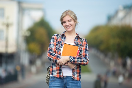 Portrait of a young attractive student girl against the blurred city street, holding copybooks, looking at the camera. Back to school concept photoの写真素材