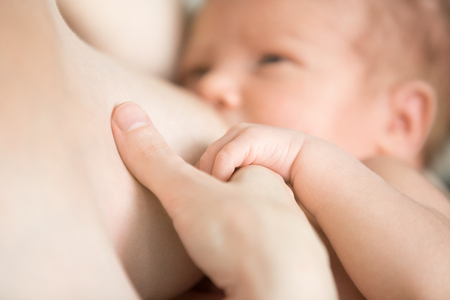 Close-up portrait of sweet newborn baby breastfeeding. Adorable new born caucaian kid holding moms finger with his cute little palm while breast feeding. Focus on hands. Young mother nursing babyの写真素材
