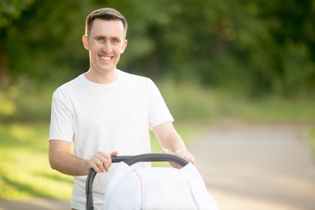 Portrait of happy young father standing in park with baby-carrier on summer day looking at camera and smiling happilyの写真素材