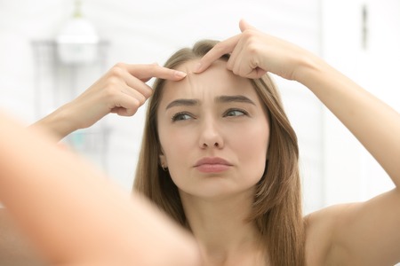 Young woman worried checking acnes on her forehead, looking at the bathroom mirror. View over the shoulder. Beauty, skin care concept, lifestyleの写真素材