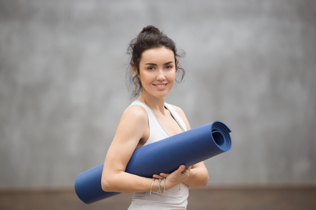 Portrait of attractive woman wearing white sportswear holding blue yoga or fitness mat after working out at home or in club. Friendly smiling sport instructor looking at camera. Healthy life conceptの写真素材