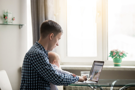 Young man working at the desk at home with laptop, holding a baby at knees, working remotely, spending time with children, stay-at-home dad, meeting computer from birth, searching a babycare centerの写真素材