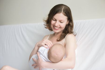Portrait of a smiling young woman holding and breastfeeding cute baby, sitting comfortably, physical and emotional health of mother and child, time to relax quietly with newborn, enjoyingの写真素材