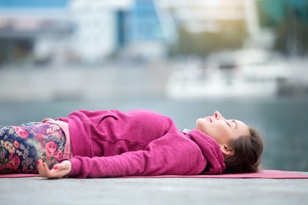 Young attractive woman practicing yoga, lying in Dead Body exercise, Savasana, Corpse pose, relaxing, refreshing after sport, wearing nice pink sportswear, outdoor city, river background, close upの写真素材