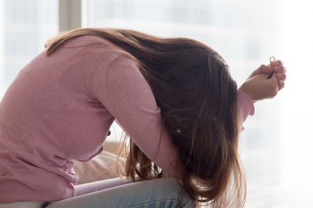 Upset depressed young woman holding wedding ring indoors, breaking off engagement, ending relationship, abandoned wife, cheated bride making hard decision, broken heart, breakup and divorce conceptの写真素材