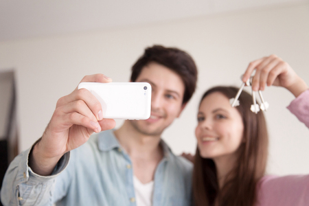 Young happy man and woman make selfie on smartphone, holding keys of new home after buying house apartment. Wife and husband purchased property, capturing moment on photo showing keys, phone in focusの写真素材