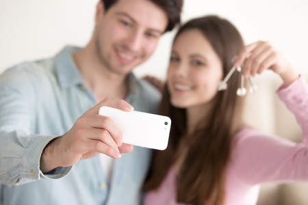 Happy smiling young couple showing keys of their new home and taking selfie picture to remember starting new life in own apartment, satisfied glad house owners with keys making photo on mobile phoneの写真素材