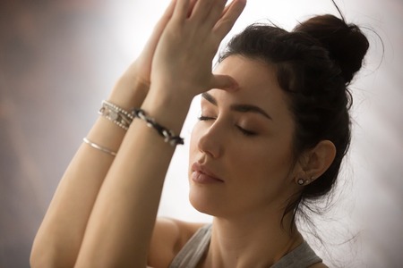Young attractive yogi woman practicing yoga concept, doing namaste gesture, namaste hands to forehead with her eyes closed, working out, wearing wrist bracelets, studio background, closeup portraitの写真素材