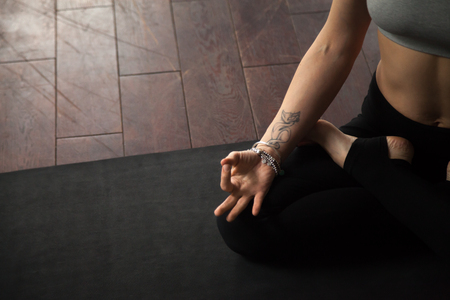 Young yogi woman practicing yoga concept, sitting on black mat in studio or home in Padmasana exercise, Lotus pose, working out, meditating, close up of hand with tattoo, copy space floor backgroundの写真素材