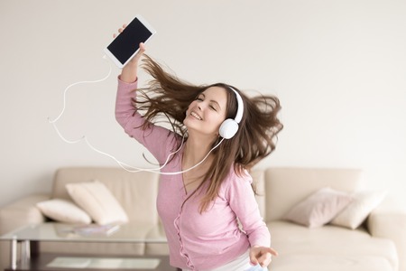 Happy girl listening to music online or radio  via tablet in headphones, teenager music lover enjoying new tracks and songs at home, young lady wearing headset dancing alone, flying hairの写真素材