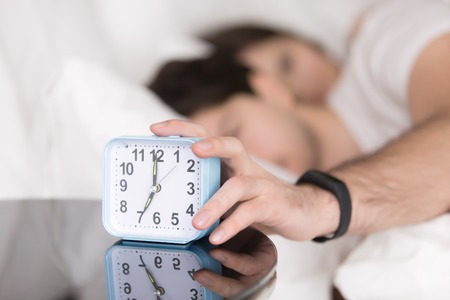 Young couple awaking after wake-up signal in the morning at home. Sleepy man stretching hand to alarm clock touching button to snooze or turn it off. Focus on hand with clock. Time to wake upの写真素材