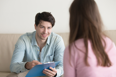 Young smiling man sitting on couch opposite woman, holding clipboard, talking with patient or asking personal information. Visiting psychologist, filling up form or questionnaire, informal interviewの写真素材