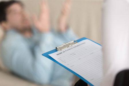 Young man on reception at the psychotherapist. Focus on clipboard with medical patient card in doctor hand. Filling personal information about new patient in clinic. Healthcare and treatment. Close upの写真素材