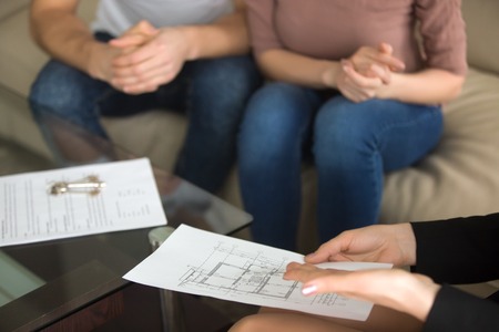 Close up view of female realtor showing to young couple of buyers flat plan on the sheet of paper sitting on sofa indoors, discussing buying apartment, considering flat purchase, interior layoutの写真素材