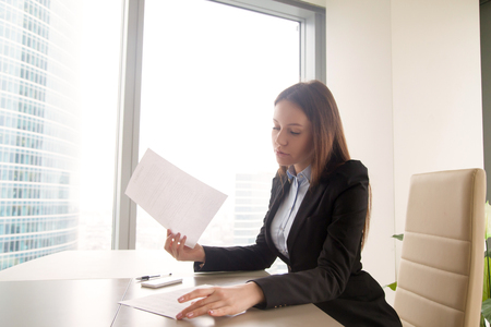 Attractive young woman wearing formal suit sitting alone at the office desk, holding some papers, reading agreements or contracts, female boss studying resume of job applicant at workplaceの写真素材
