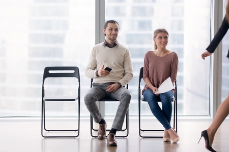 Confident businesswoman walking by male and female job candidates sitting on chairs, following her...