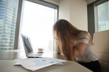 Exhausted businesswoman with disheveled hair sitting at the table and holding head in hands. Depressed female office worker in despair. Woman feeling fatigue, loosing productivity because of overworkの写真素材