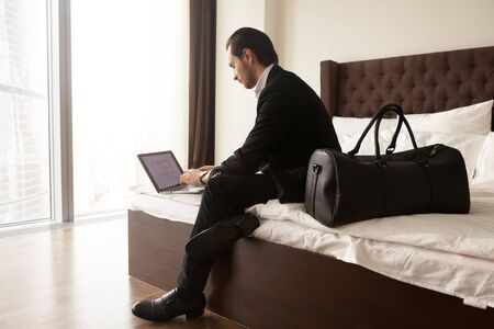 Real estate agent examines house drawing on laptop screen before travel flight. Businessman tenant arrived in hotel with luggage and working on laptop. Guy checks presentation for meeting with partnerの写真素材