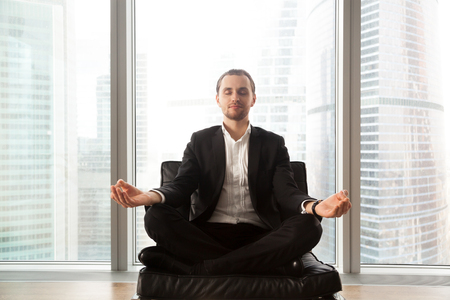 Young businessman sitting in soft chair in lotus position. Calm man in business suit meditating on couch with crossed legs. Entrepreneur engage in spiritual practices for mental health, stress reliefの写真素材