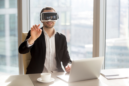 Businessman enjoys digital simulation with virtual reality glasses. Man with VR headset on head touching virtual objects. Entrepreneur working in virtual office application while sitting at workplaceの写真素材