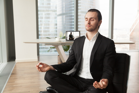Businessman meditates in lotus position on coach in modern office. Company leader practicing yoga during break at work for strength recovery and stress relief. Man relaxing near workplace with laptopの写真素材