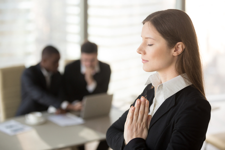 Young beautiful businesswoman enjoys meditating standing in office with eyes closed, put hands together as in prayer, reduce stress, relief to keep calm, positive thinking, appeasement, autosuggestionの写真素材